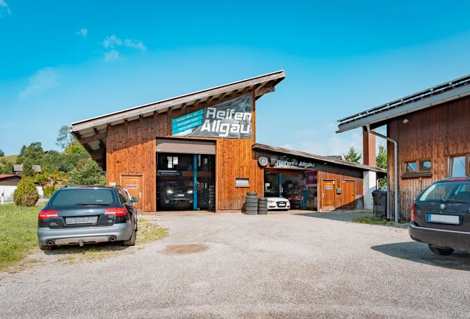 Holzverkleidete Werkstatthalle von Reifen Allgäu mit geöffnetem Hallentor und Kundenautos auf dem Kiesplatz davor