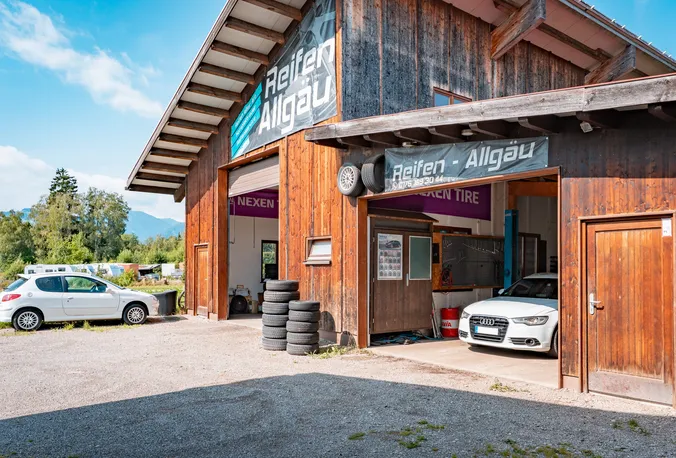 Betriebsgebäude von Reifen Allgäu mit zwei offenen Toren, Reifenstapeln davor und den Allgäuer Bergen im Hintergrund